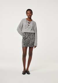 Grauer Pullover mit Schnürung vorne, kombiniert mit einem schwarzen gemusterten Minirock. Das Outfit wird mit schwarzen, detaillierten Ballerinas abgerundet.