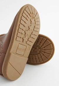 Zapatos de cuero marrón con una suela de goma texturizada en tono beige que presenta un patrón de estrellas, puntera redondeada y detalle de logo en el talón.