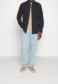 Conjunto informal que incluye una camisa de botones navy sobre una camiseta beige, jeans azul claro y zapatillas marrón claro con detalles en blanco.