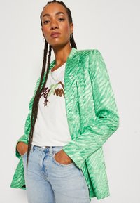 Sacou texturat verde cu un finisaj satinat, dotat cu închidere cu un singur buton și rever cu tăietură, asortat cu un tricou alb brodat și blugi.