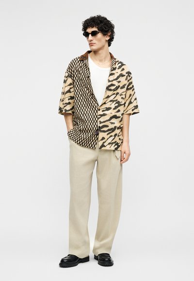 Man met zwarte zonnebril, half geruit en half dierenprint korte mouwen overhemd, witte ondershirt, beige broek en zwarte schoenen.