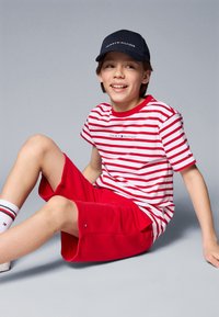 Rød og hvidstribet bomulds T-shirt, røde shorts, sort kasket med logo, hvide sokker med røde detaljer, siddende mod en grå baggrund.