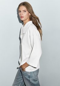 Weiße übergroße Bluse mit einem Notch-Kragen und weiten Ärmeln, kombiniert mit hellen Denim-Jeans. Glatte Textur, minimalistisches Design.