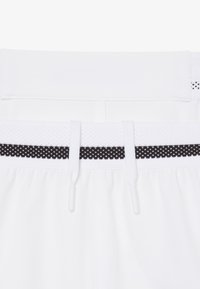 Vita elastica bianca con striscia puntinata nera e cordini bianchi su tessuto bianco, dettaglio ravvicinato di un indumento.