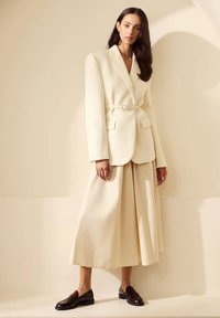 Crèmekleurige blazer met scherpe revers en een ceintuur, gecombineerd met crème kleurige wijde broek en donkere instapschoenen, tegen een neutrale achtergrond.