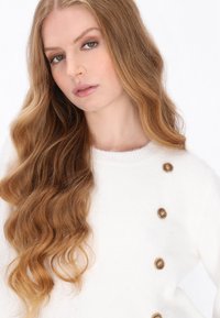 Hvid sweater lavet af blødt materiale med en rund halsudskæring, sideknapper med trædekorationer, og langt bølget hår, der falder ned.