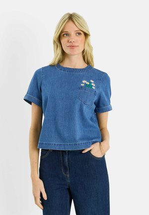 Mujer rubia que lleva un top corto de mezclilla azul con un diseño floral en el bolsillo y vaqueros azul oscuro, con la mano descansando en el bolsillo.