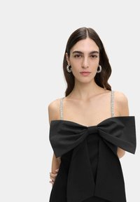 Černé šaty s velkou mašlí vpředu, ozdobnými špagetovými ramínky, měkkou tkaninou a strukturovaným designem. Třpytivé detaily na ramínkách.