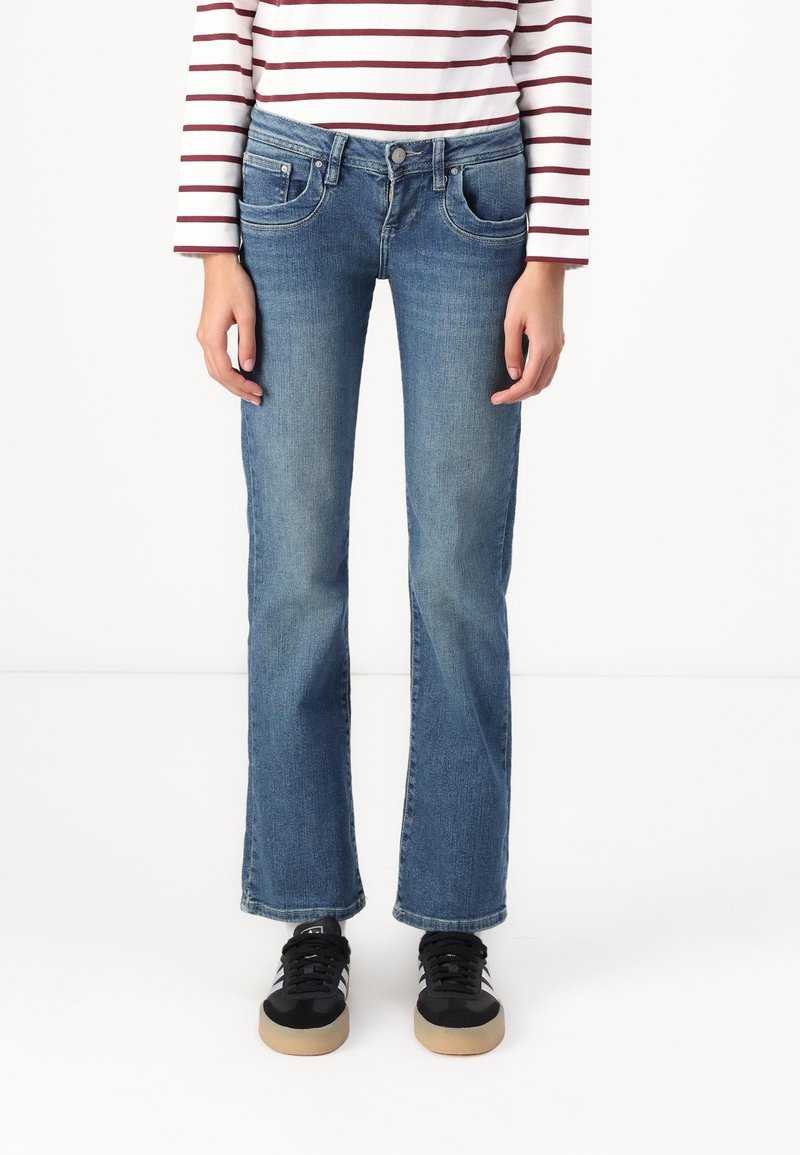 Jeans de mezclilla azul con corte recto, diseño de cinco bolsillos y un ligero desgaste. Combinados con zapatillas negras que tienen una suela de goma.