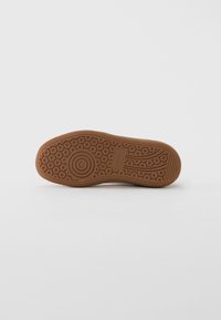 Brun gummisula på en Puma-sneaker som visar cirkulära och linjära slitbanemönster mot en blank vit bakgrund.