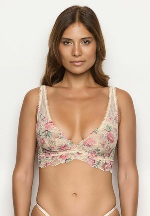 Donna con capelli castano di media lunghezza, che indossa un bralette in pizzo floreale beige e un completino coordinato, in piedi contro uno sfondo chiaro uniforme.