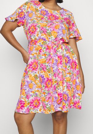 Nainen, joka pukeutunut lyhythihaiseen, polvipituiseen mekkoon, jossa on vaaleanpunainen, oranssi ja violetti kukkakuosi ja kangasvyö solmittuna vyötärölle.