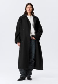 Dlhý čierny vlnený kabát s dvojradovým zapínaním, výstrihom s výrezmi, širokými rukávmi a nadrozmerným strihom, nosený cez svetlý sveter a tmavé nohavice.