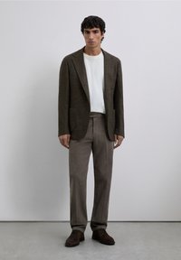 Blazer marrón texturizado sobre una camiseta blanca, combinado con pantalones marrones claros y zapatos marrones oscuros, de pie contra un fondo neutro.