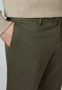 Personne portant un pantalon vert olive et un pull en maille beige, avec la main gauche partiellement dans la poche.