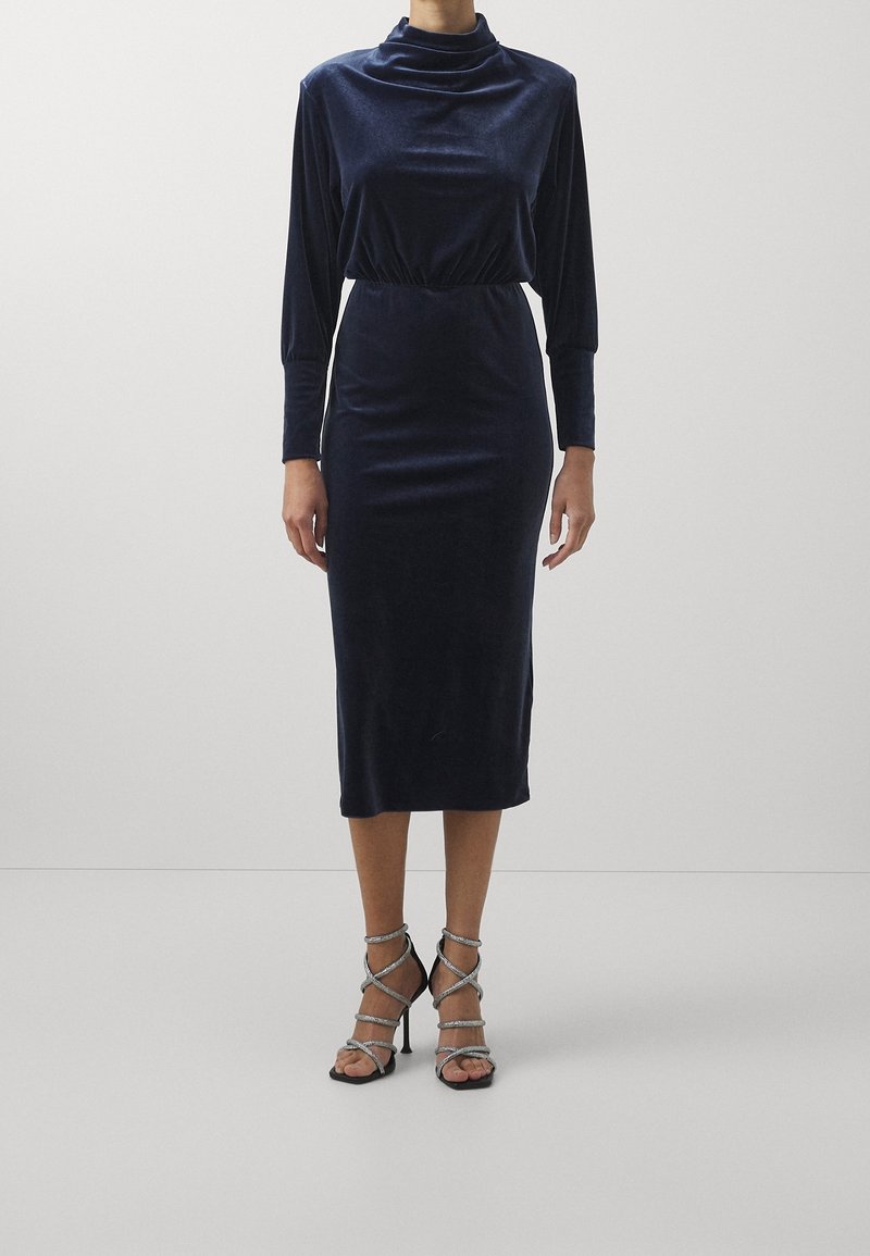 Femme portant une robe midi en velours bleu marine à manches longues avec un col montant et des talons hauts argentés à brides.