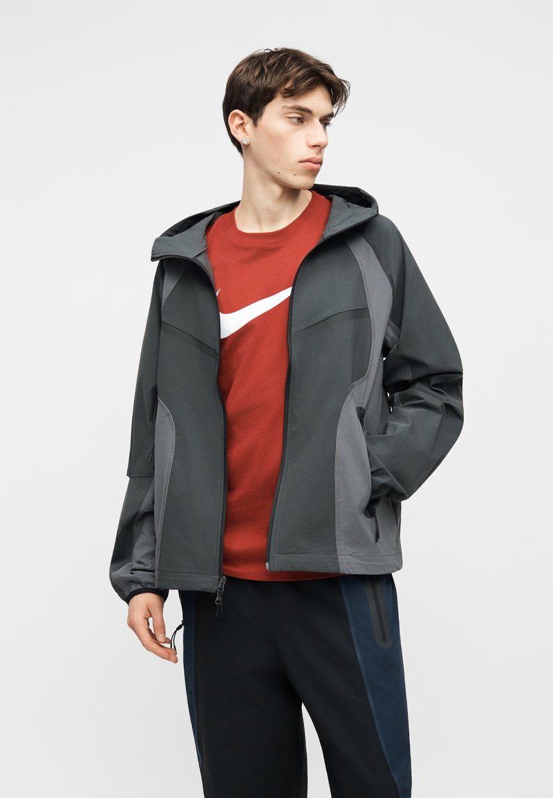 Junger Mann trägt eine dunkelgraue Kapuzenjacke über einem roten Nike T-Shirt und schwarzen Hosen, schaut nach links und hat die Hände in den Taschen.