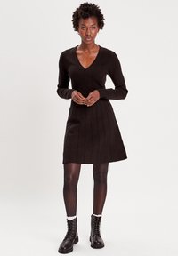 Schwarzes geripptes Kleid mit V-Ausschnitt und langen Ärmeln, das einen enganliegenden Oberkörper und einen ausgestellten Rock hat, kombiniert mit schwarzen Schnürstiefeln.