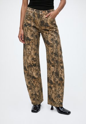 Persoon die een broek met hoge taille en brede pijpen draagt met een bruin-zwart abstract patroon, een zwarte top en zwarte hakken, staand met één hand in de zak.