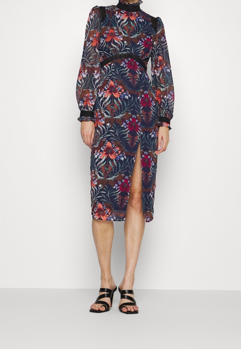 Šaty s květinovým potiskem, dlouhými rukávy, vysokým výstřihem a rozparkem po straně. Mají tmavé pozadí s barevnými květy a texturovanými manžetami.