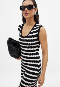 Robe en tricot à rayures noir et blanc avec un col rond, assortie de lunettes de soleil noires et d'une pochette tissée noire. Cheveux longs et lisses.