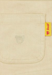 Bolsa de tela beige con un logotipo de oso en relieve. Etiqueta lateral amarilla con texto en rojo, que indica el nombre de la marca, ubicada a la derecha.