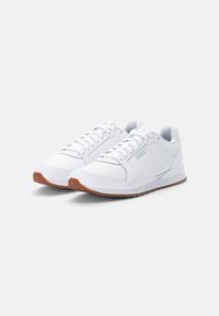 Bílé sportovní tenisky s hladkým syntetickým svrškem, s šněrovacím designem, polstrovaným límcem a hnědou gumovou podrážkou. Minimalistické logo.