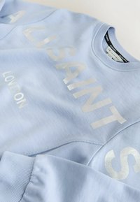 Šviesiai mėlynas džemperis su blizgiais sidabriniais užrašais. Turi ribinuotas rankogalius ir kaklo iškirptę, ryškiai užrašyti "ALLSAINTS" ir "LOVDON".