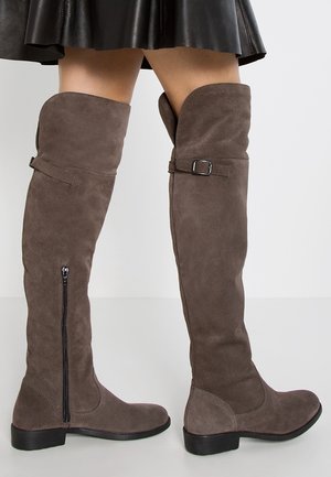 Ruskeat mokkanahkaiset polven yläpuolelle ulottuvat saappaat, joissa on sivuvetoketju ja solkikoriste, yhdistetty mustaan nahkahameeseen yksinkertaisen taustan edessä.