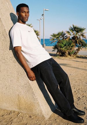 Mann in weißem T-Shirt und schwarzer Hose lehnt an einer Betonstruktur am Sandstrand mit Palmen und Ozean im Hintergrund.