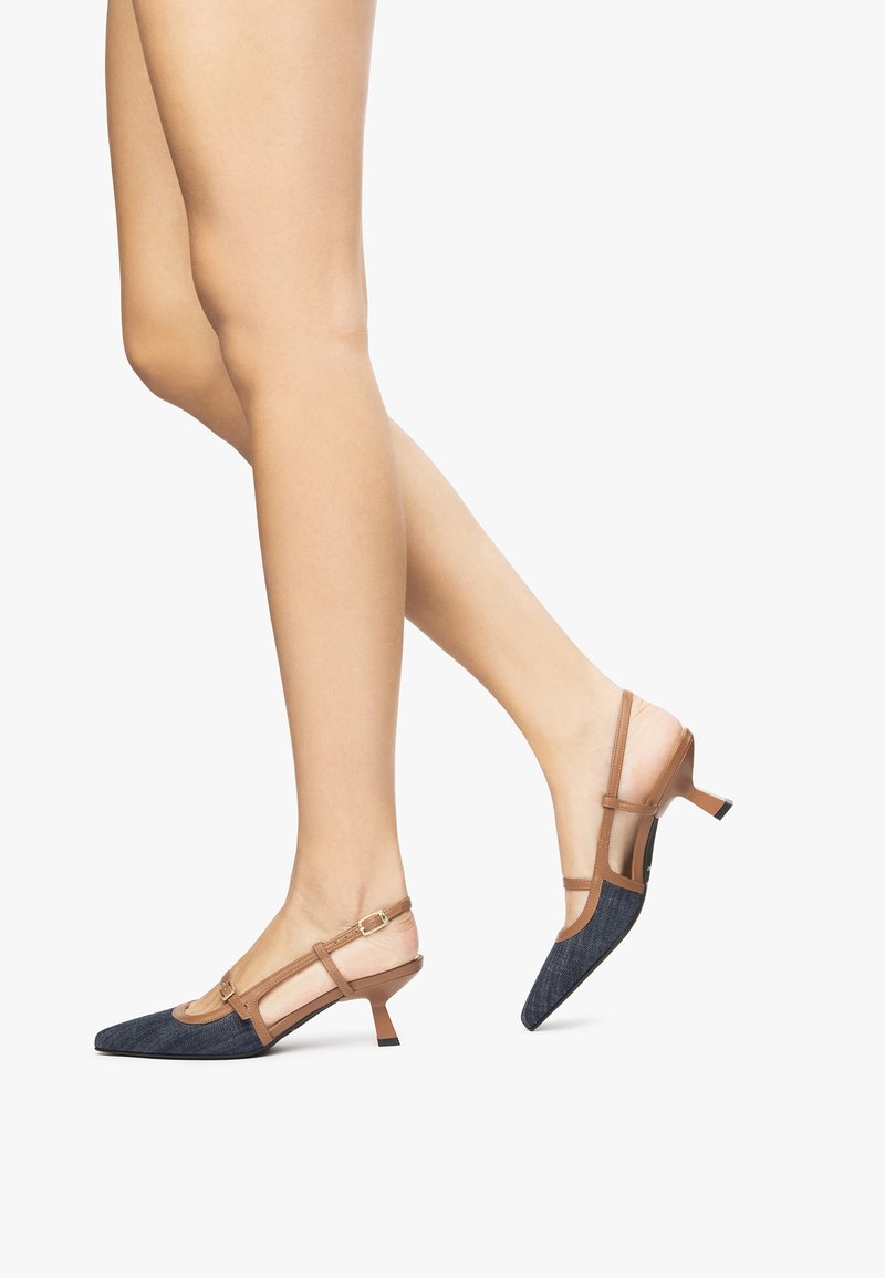 Jambes portant des escarpins slingback à bout pointu avec devant en tissu bleu et brides en cuir marron clair, et petits talons aiguilles, sur fond blanc.