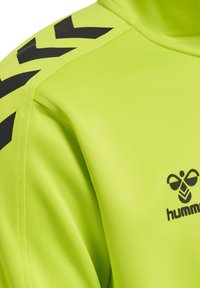 Jasnożółto-zielony sportowy top z czarnymi ukośnymi paskami na ramieniu i czarnym logo pszczoły z napisem „hummel” na piersi.