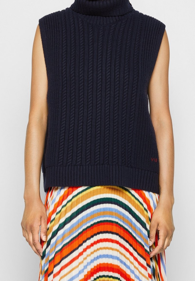 Suéter sin mangas de cuello alto azul marino con patrón de punto trenzado, combinado con una falda plisada a rayas multicolores.