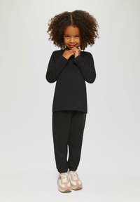 Jong kind met krullend haar, gekleed in een zwarte lange mouw top en broek, staande met handen dicht bij de kin, glimlachend, tegen een lichte effen achtergrond.