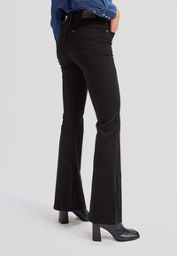 Personne portant un jean flare noir taille haute, des bottes noires à talons et une chemise bleue à manches longues, debout avec une main dans la poche.