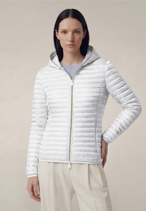 Kobieta z prostymi ciemnymi włosami, ubrana w białą pikowaną kurtkę z kapturem i zamkiem oraz beżowe spodnie, stojąca z jedną ręką na biodrze.