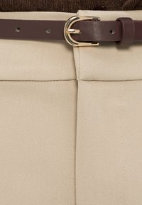 Ceinture en cuir marron avec une boucle dorée, attachée à un pantalon beige taille haute, assortie à un haut marron à texture.