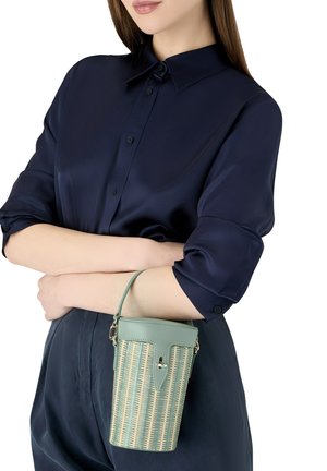 Vrouw in een marineblauw overhemd met opgerolde mouwen, die een kleine groene geweven emmerzak met leren riem en sluiting vasthoudt.