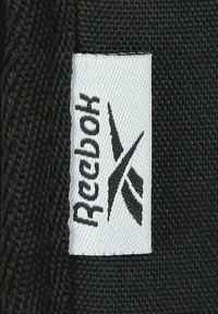 Etiqueta de tela blanca con el logo y texto negro de Reebok cosidos sobre un material texturizado negro.