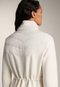 Jachetă din fleece alb cu guler înalt, având un design cojocat în formă de zigzag pe umeri și un detaliu cu talie strânsă.