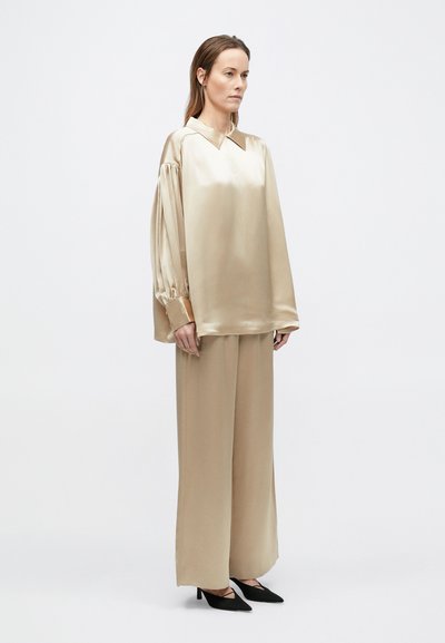 Žena stojí oblečená v voľnej krémovej saténovej blúzke s dlhými nafúknutými rukávmi a širokými nohavicami, doplnená o čierne lodičky so zašpicatou špičkou.