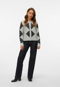 Grijze cardigan met zwarte diamantpatronen, ronde halslijn, knoopsluiting, gecombineerd met donkere spijkerbroek en zwarte hoge laarzen.