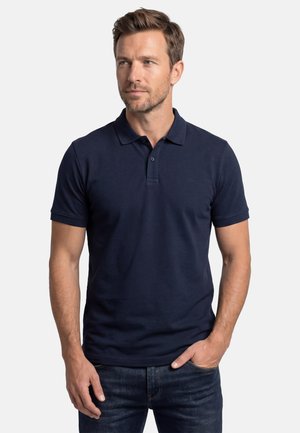 Mand med lysebrunt hår og skægstubbe iført en navyblå polo med korte ærmer og mørke jeans, stående med den ene hånd i lommen.