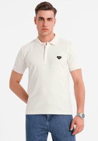 Bílá polo košile s knoflíkovým límcem, s malým černým trojúhelníkovým logem na levé části hrudi. Krátké rukávy a měkká textura tkaniny.