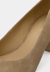 Beige ruskindpumpe med rundet tå og en mellemhøj blokhæl. Indersiden har en glat beige foring med diskrete syningsdetaljer.