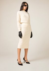 Vestito color crema con strass argento, maniche lunghe e collo alto; abbinato a guanti in pelle nera e décolleté nere con tacco.