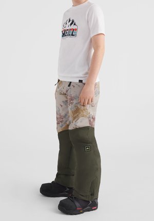Person iført en hvid grafisk t-shirt og mønstrede bukser med mørkegrønne beskyttelsesgamacher og sorte udendørs sko.