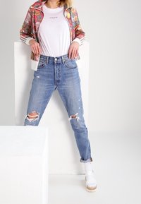 Biała koszulka pod wielokolorową kurtką z zamkiem błyskawicznym, zestawiona z jasnoniebieskimi, podartymi dżinsami i białymi sandałami na koturnie.