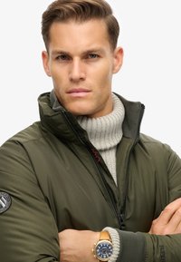 Olivgrüne Daunenjacke mit hohem Kragen, Reißverschluss und Logopatch; über einem hellgrauen gestreiften Rollkragenpullover getragen; goldene Uhr mit blauem Ziffernblatt.