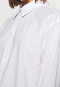 Chemise blanche à boutons avec col classique, tissu en coton lisse et coupe ample. Comprend une patte de boutonnage complète et des manches longues.